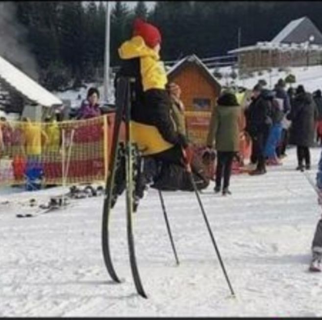 Eine Person sitzt auf Skistöcken, die an Skiern befestigt sind, auf einer Skipiste. Im Hintergrund sind weitere Menschen und Gebäude zu sehen.