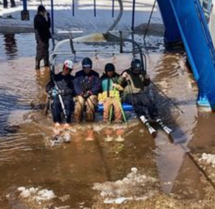 Eine Gruppe von vier Skifahrern sitzt auf einem Sessellift, der sich über eine überflutete Piste bewegt. Die Skifahrer tragen Skikleidung und wirken unbeeindruckt.