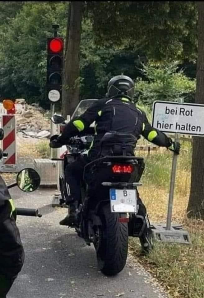 Ein Motorradfahrer hält ein "Bei Rot hier halten" Schild an einer Baustelle, während die Ampel rot zeigt. Sein Motorrad hat ein Berliner Kennzeichen.