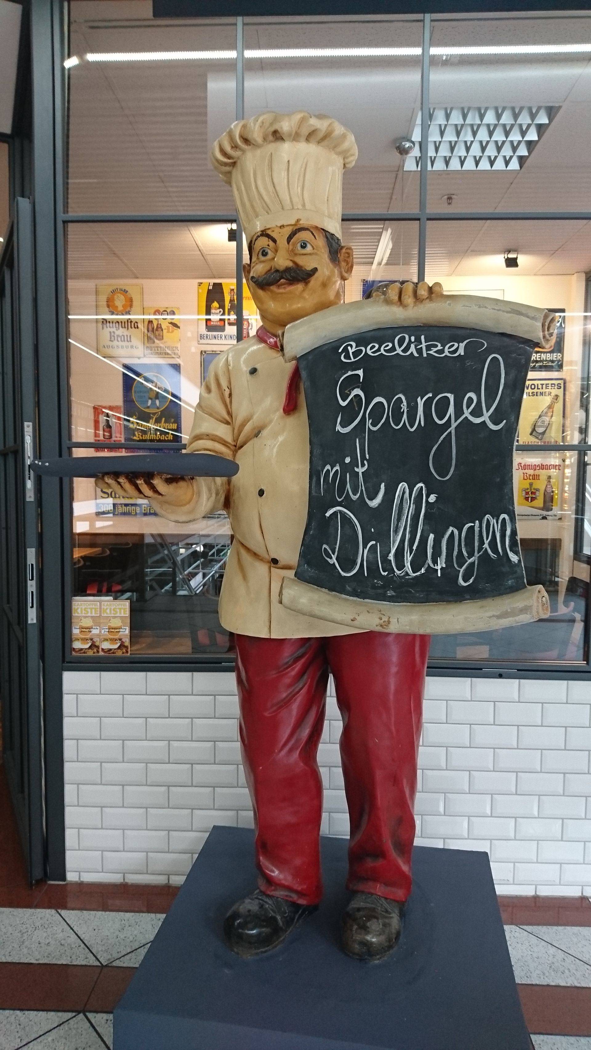 Eine Statue eines Kochs mit einem großen Schnurrbart und Kochmütze hält eine Tafel. Auf der Tafel steht "Beelitzer Spargel mit Drillingen".