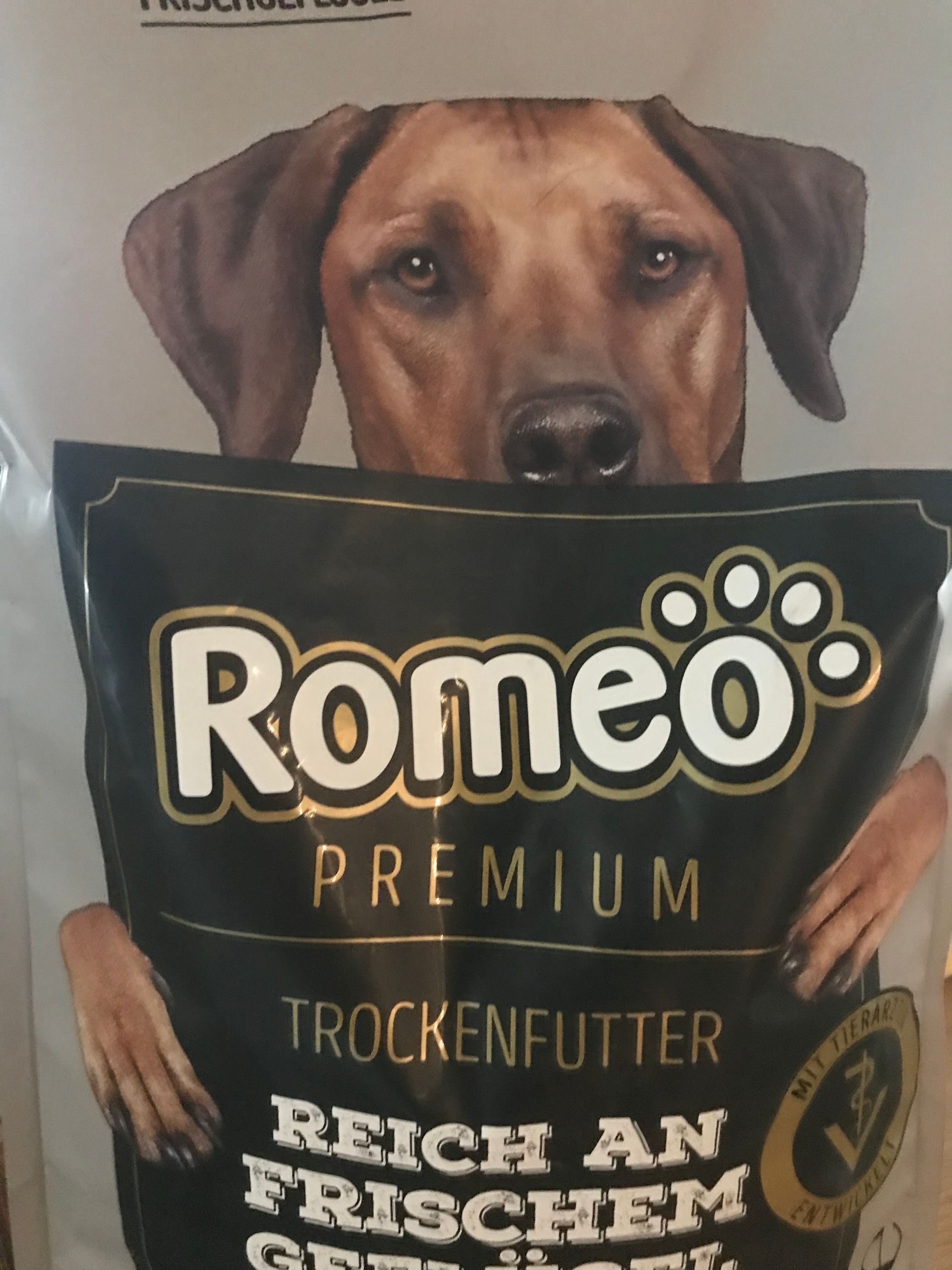 Ein Hund schaut über eine Tüte Hundefutter der Marke "Romeo" mit der Aufschrift "Premium Trockenfutter, reich an frischem Geflügel". Die Tüte wird von zwei Händen mit roten Nägeln gehalten.