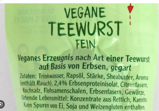 Das Bild zeigt eine Nahaufnahme der Verpackung einer "Veganen Teewurst". Auf der Packung steht in großer Schrift "VEGANE TEEWURST FEIN". Darunter folgt der Text "Veganes Erzeugnis nach Art einer Teewurst auf Basis von Erbsen, gegart." Die restlichen Zutaten und Warnhinweise sind in kleinerer Schrift unterhalb aufgeführt.