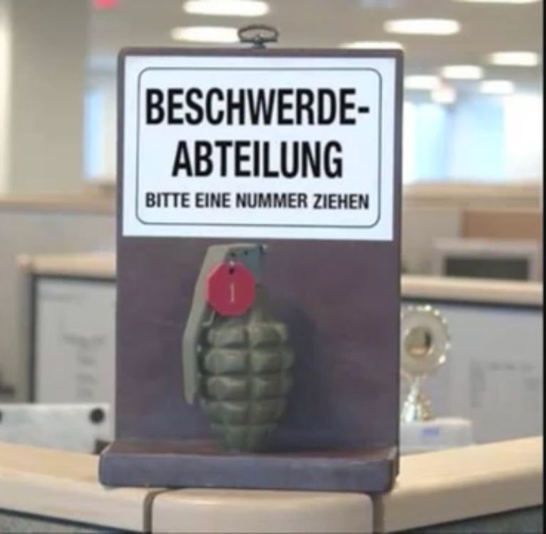 Eine Granate mit der Nummer 1 hängt vor einem Schild mit der Aufschrift "BESCHWERDE-ABTEILUNG. BITTE EINE NUMMER ZIEHEN."