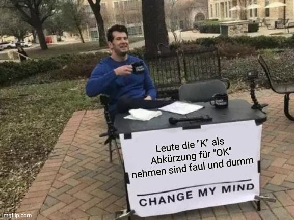 Ein Mann sitzt an einem Tisch im Freien und hat ein Schild vor sich stehen auf dem steht: "Leute die "K" als Abkürzung für "OK" nehmen sind faul und dumm. CHANGE MY MIND"