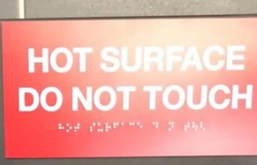 Ein rotes Schild mit der Aufschrift "HOT SURFACE DO NOT TOUCH" in weißer Schrift. Unter der Schrift befindet sich ein Text in Braille.