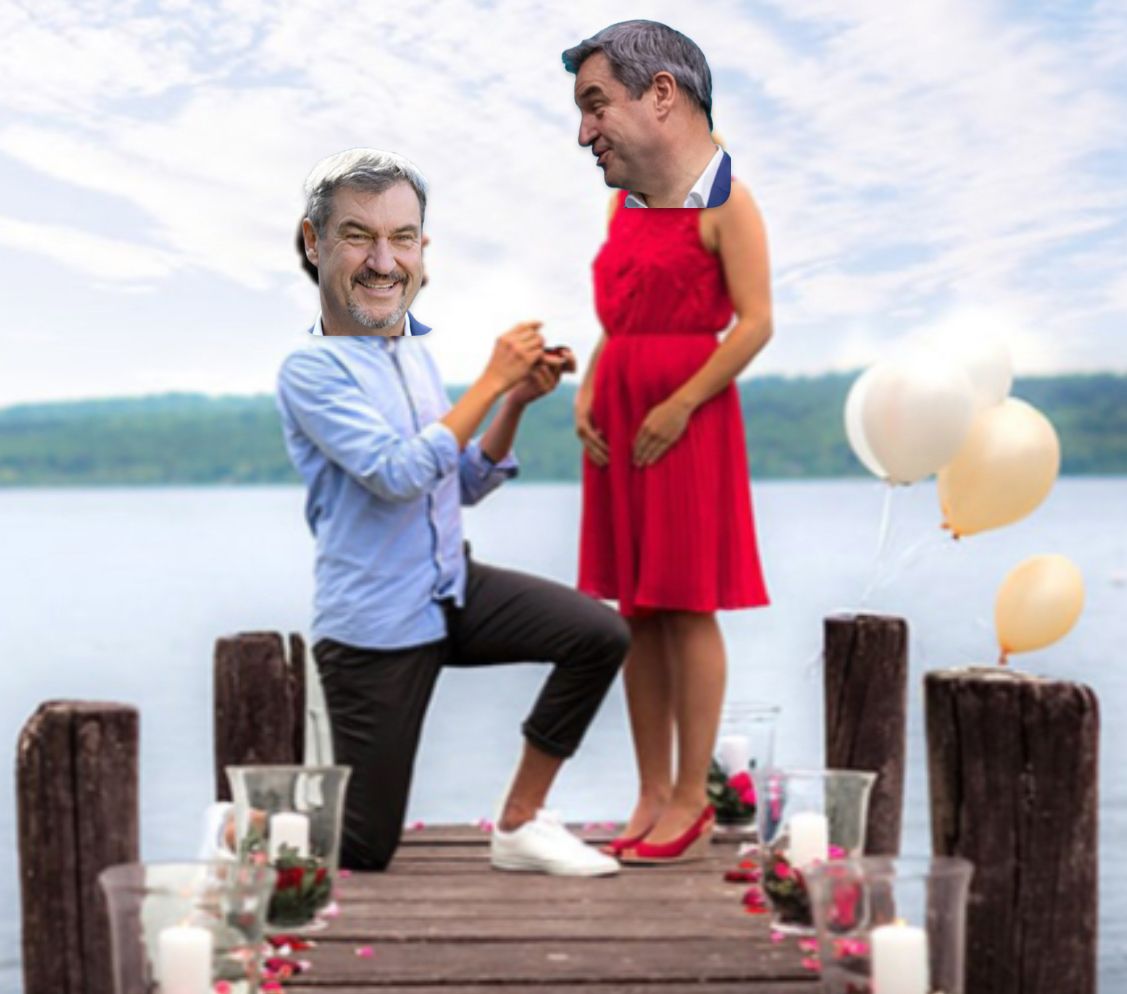 Ein Bild zeigt Markus Söder, wie er auf einem Steg vor einem See kniet und sich selbst einen Heiratsantrag macht. Er trägt ein hellblaues Hemd und dunkle Hosen. Er hält eine kleine Schatulle mit einem Ring. Die Version von Markus Söder, die den Antrag annimmt, trägt ein rotes Kleid und rote Schuhe. Der Hintergrund zeigt einen See und einige weiße und orangefarbene Luftballons. Es sind einige Kerzen auf dem Steg zu sehen.