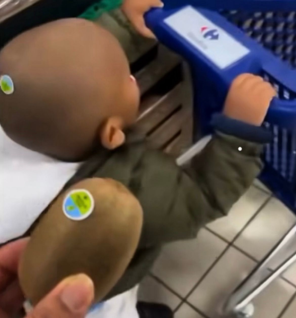 Ein Baby mit einem Aufkleber auf dem Kopf und einer Kiwi, die auch einen Aufkleber hat. Das Baby steht in einem Einkaufswagen.