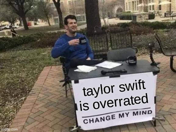 Ein Mann sitzt an einem Tisch im Freien und hält eine Tasse. Auf dem Tisch steht ein Schild mit der Aufschrift "taylor swift is overrated. CHANGE MY MIND"