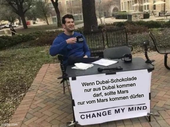 Ein Mann sitzt an einem Tisch im Freien mit einer Tasse und einem Mikrofon. Auf dem Tisch steht ein Schild mit der Aufschrift: "Wenn Dubai-Schokolade nur aus Dubai kommen darf, sollte Mars nur vom Mars kommen dürfen. CHANGE MY MIND."