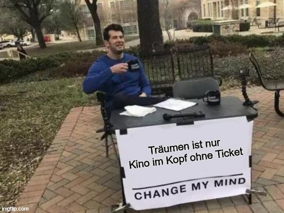 Ein Mann sitzt an einem Tisch im Freien. Er hat einen Kaffeebecher in der Hand. Auf dem Tisch steht ein Schild mit dem Text: "Träumen ist nur Kino im Kopf ohne Ticket. CHANGE MY MIND".