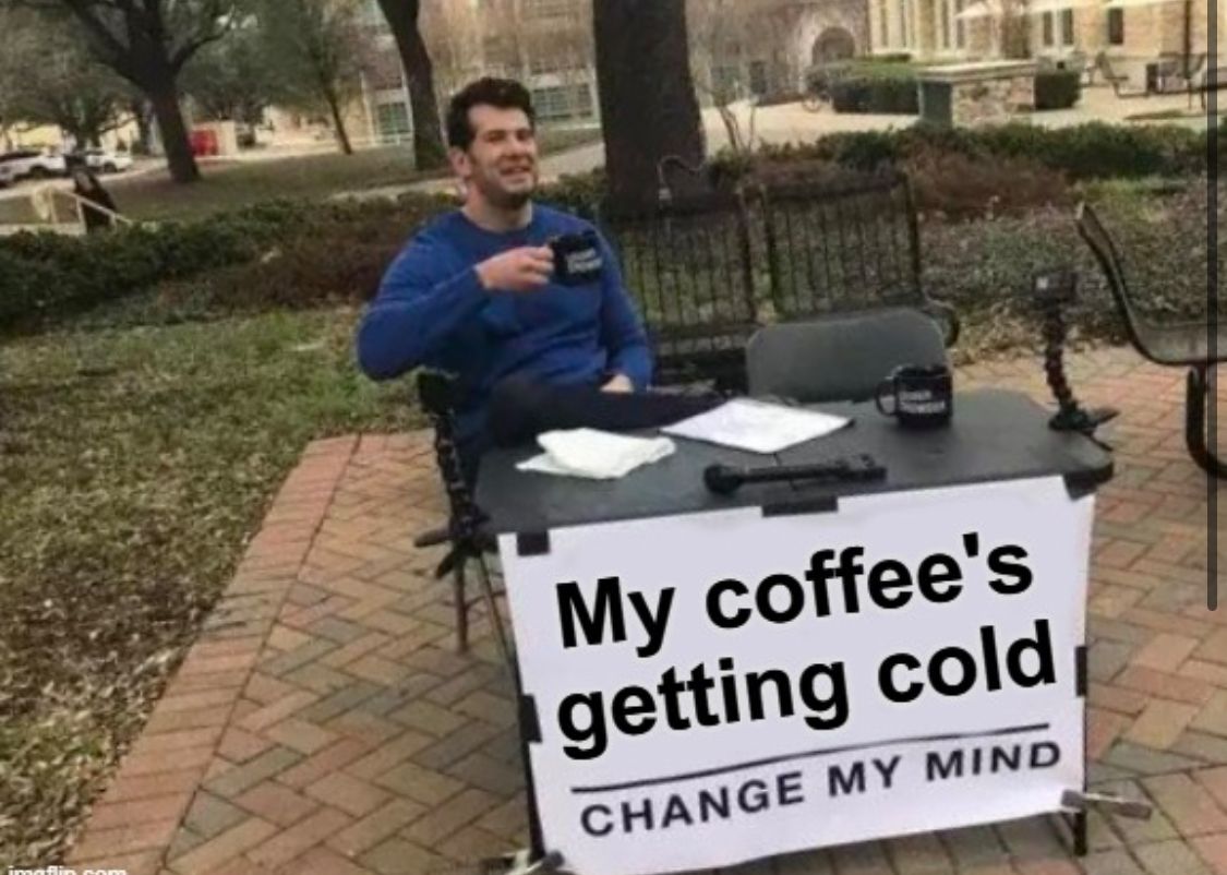 Ein Mann sitzt an einem Tisch im Freien mit einem Becher in der Hand. Vor ihm steht ein Schild mit der Aufschrift: "My coffee's getting cold. CHANGE MY MIND"